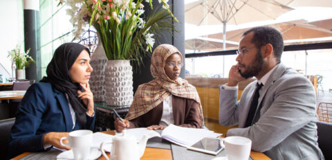 Multiethnic business partners working on agreement in cafe. Businessman and Muslim businesswomen sitting in coffee shop, calling on phone and signing documents. Partnership concept